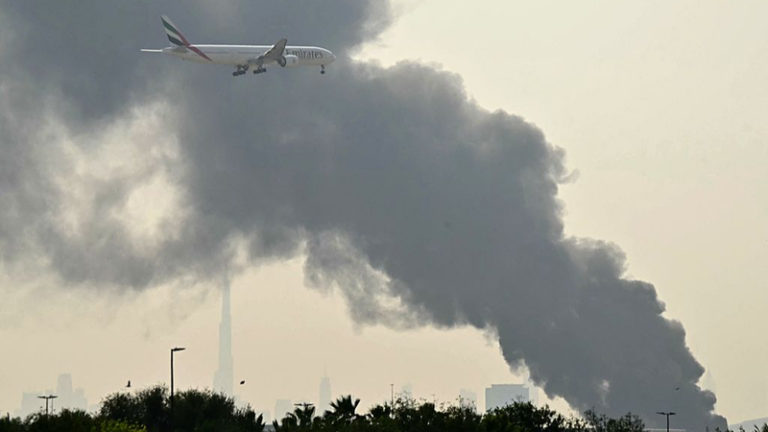 Emirates passenger jet flying above a thick plume of dark smoke rising over a city skyline.